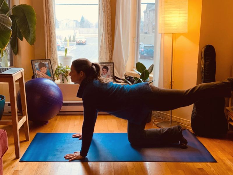 Alida on all fours on a blue mat, one leg extended, in front of a large window