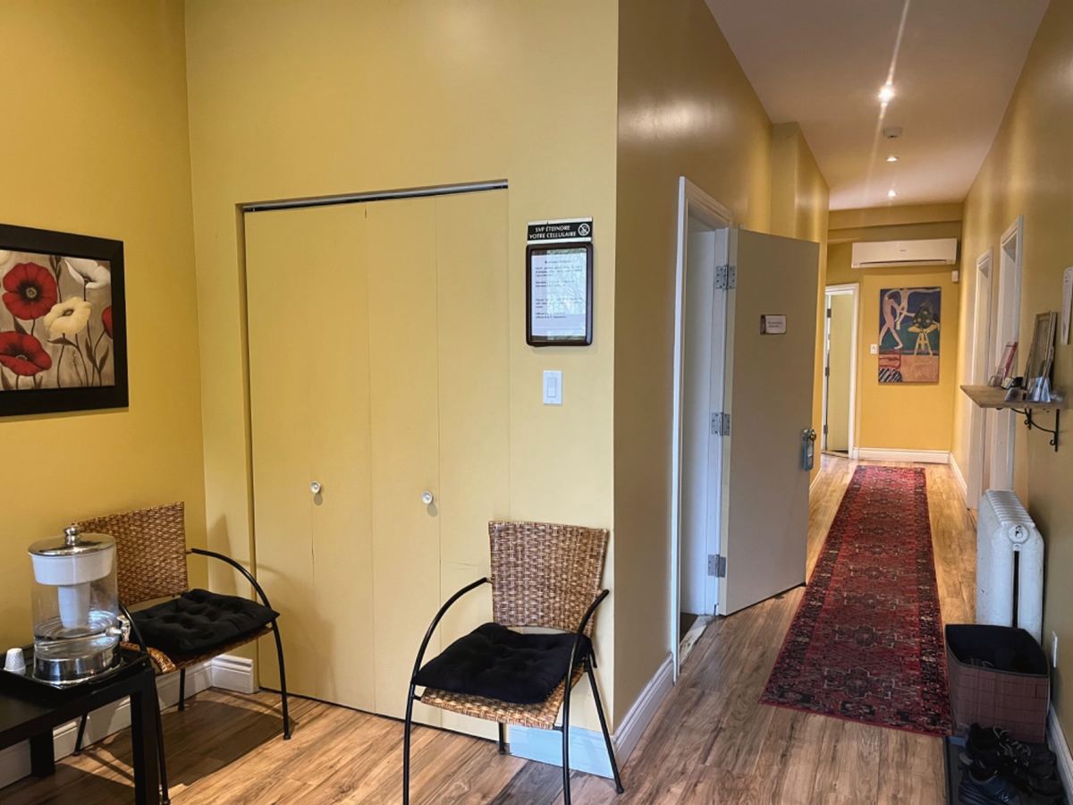 La salle d'attente de la clinique, murs jaunes, deux chaises de rotin, et le corridor au long tapis rouge
