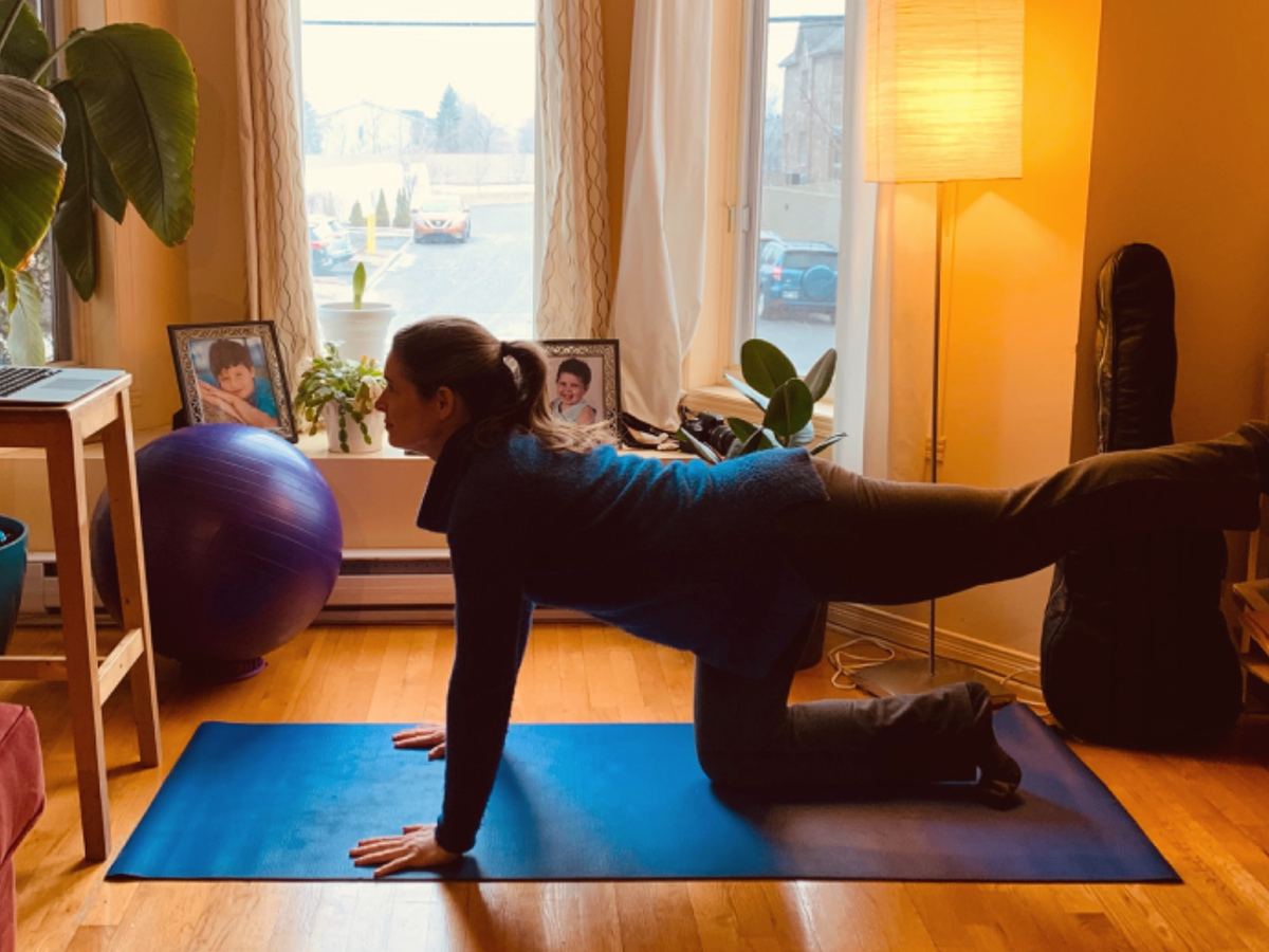 Alida Farrell on all fours on a blue mat, one leg extended, in front of a large window