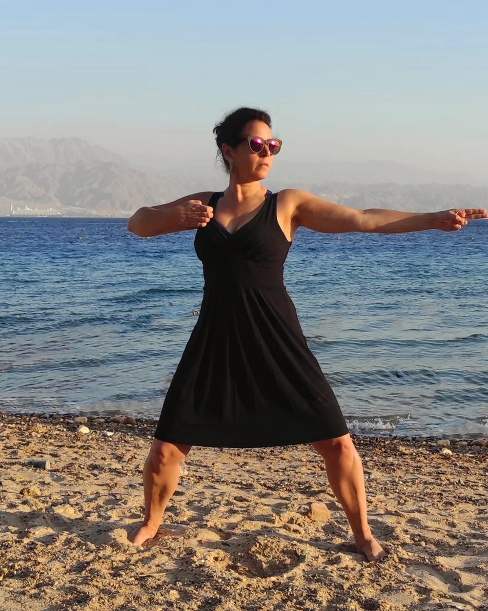 Sharonne Cohen dans une posture de Qi Gong, pieds nus sur une plage, la mer derrière